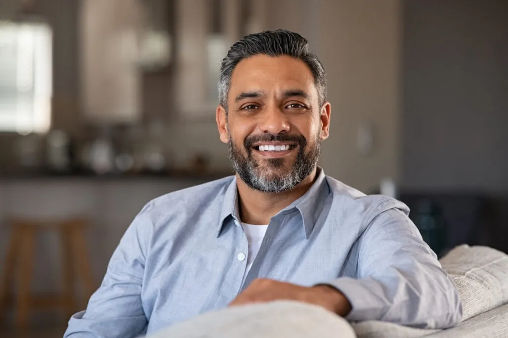 Smiling man with gray beard sits on a couch in a cozy living room, wearing a light blue shirt.