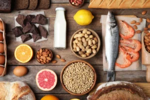 A variety of foods on a wooden table: dark chocolate, milk, lemon, fish, shrimp, peanuts, almonds, oranges, eggs, bread, and grains.