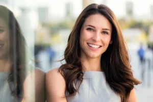 Smiling woman with long brown hair leans against glass, reflecting her image.