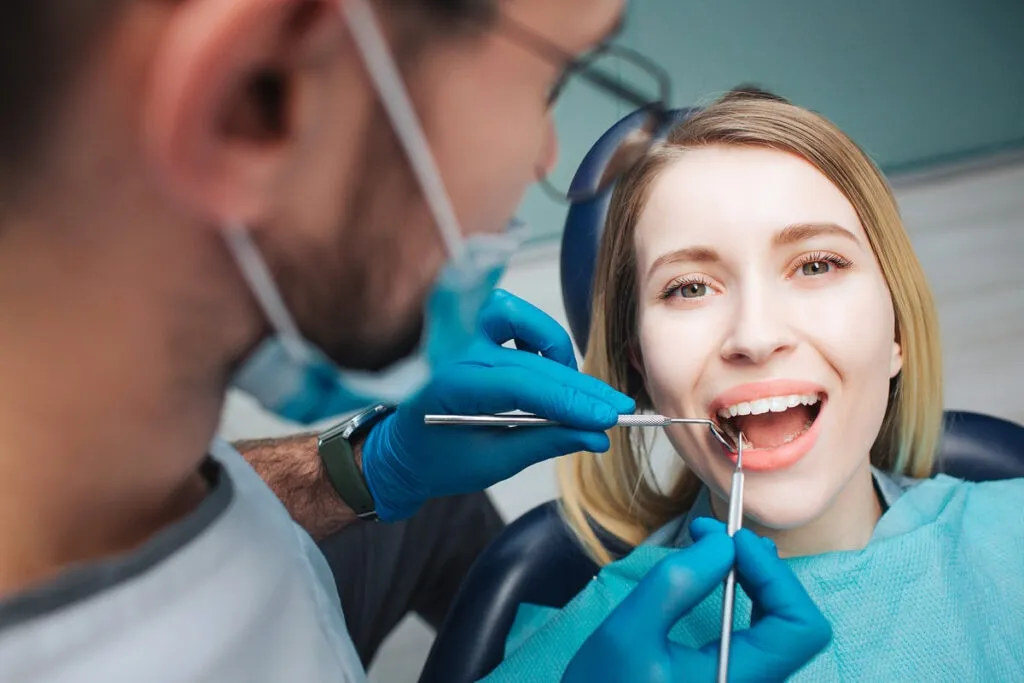 Female client lying in chair in dentistry room. She keep mouth opened and look on camera. Doctor use teeth tools for treatment