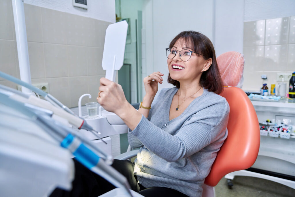 Smiling middle aged woman in dental chair in dentist's office with mirror looking at her teeth. Treatment, therapy, dental care, prosthetics, dentistry concept