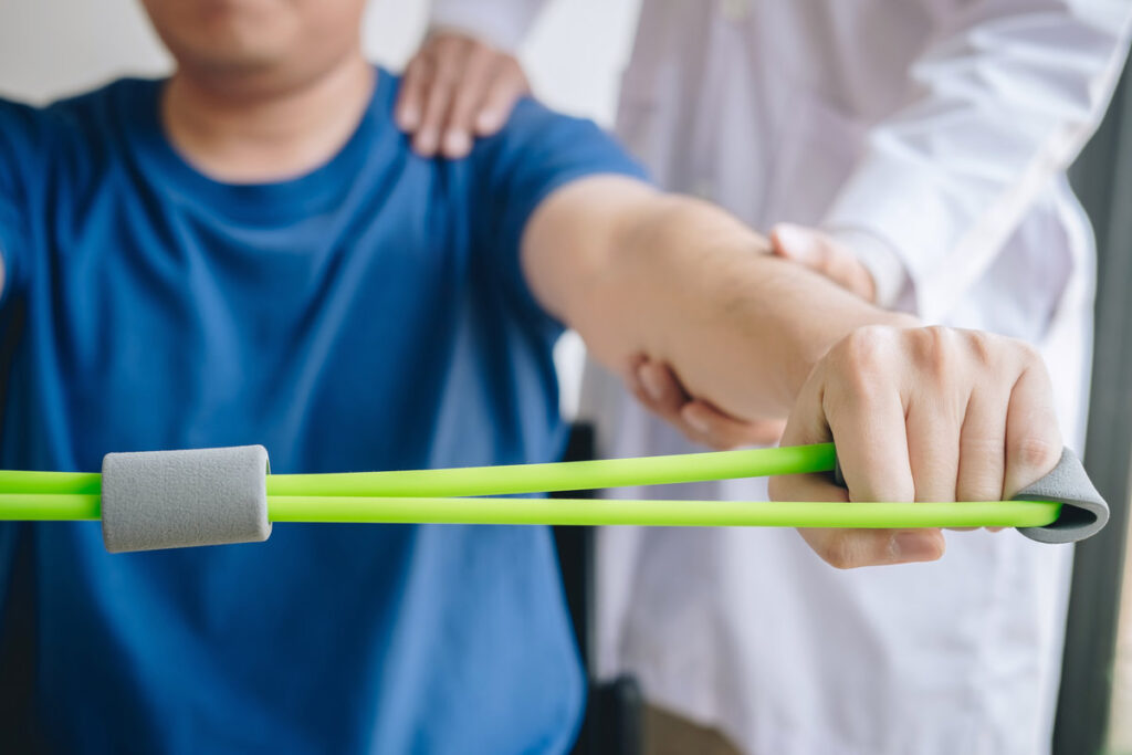 Doctor physiotherapist assisting a male patient while giving exercising treatment on stretching his arm with exercise band in the clinic, Rehabilitation physiotherapy concept.