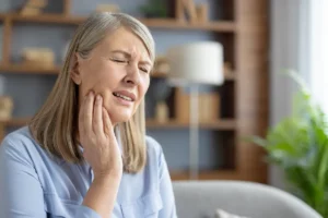 An elderly Caucasian woman sits at home, touching her cheek and grimacing in pain. She appears to be suffering from a severe toothache or jaw discomfort.